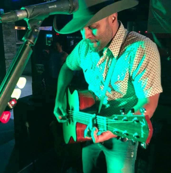Nigel Edison playing acoustic guitar in a cowboy hat under green light
